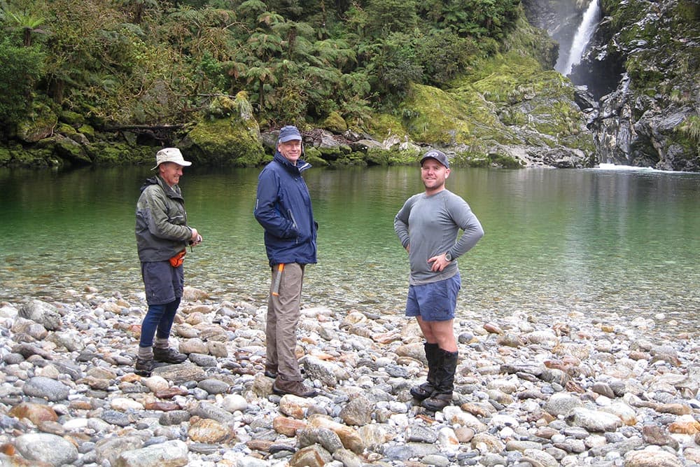 Three team members beside a river pool below a waterfall